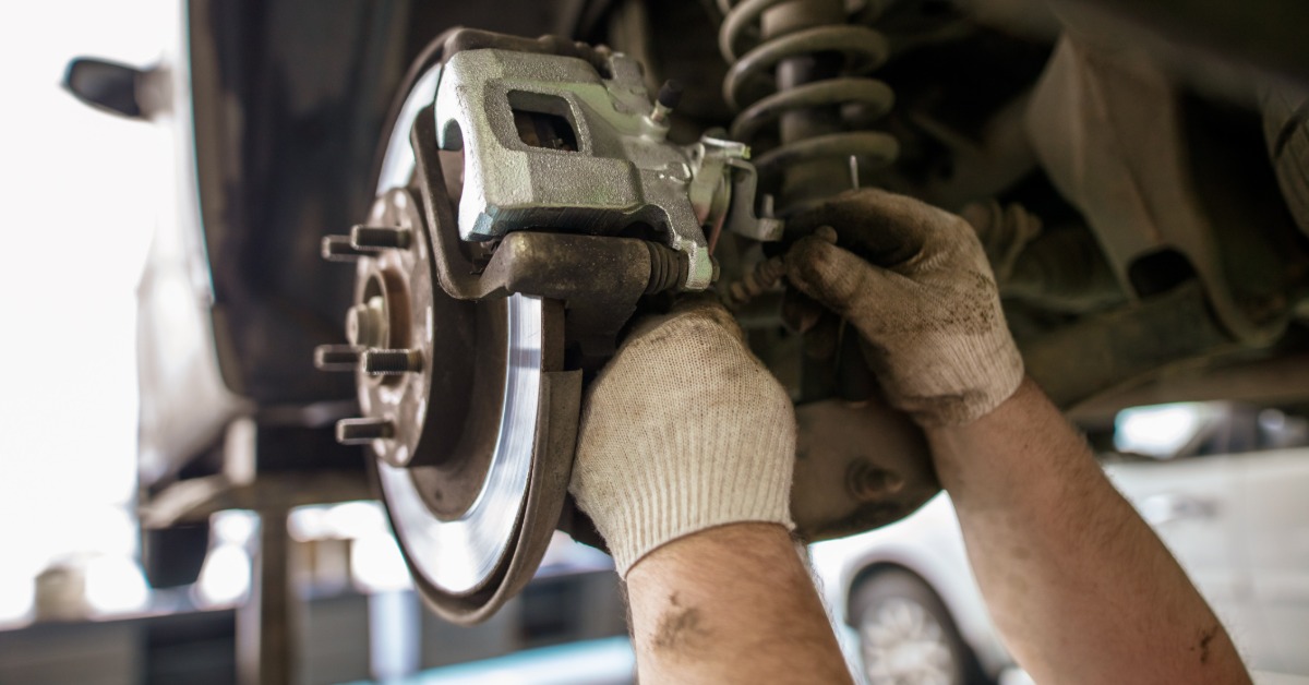 Two gloved hands are working on a car brake assembly with a silver disc, caliper, coil spring, and exposed wheel studs.