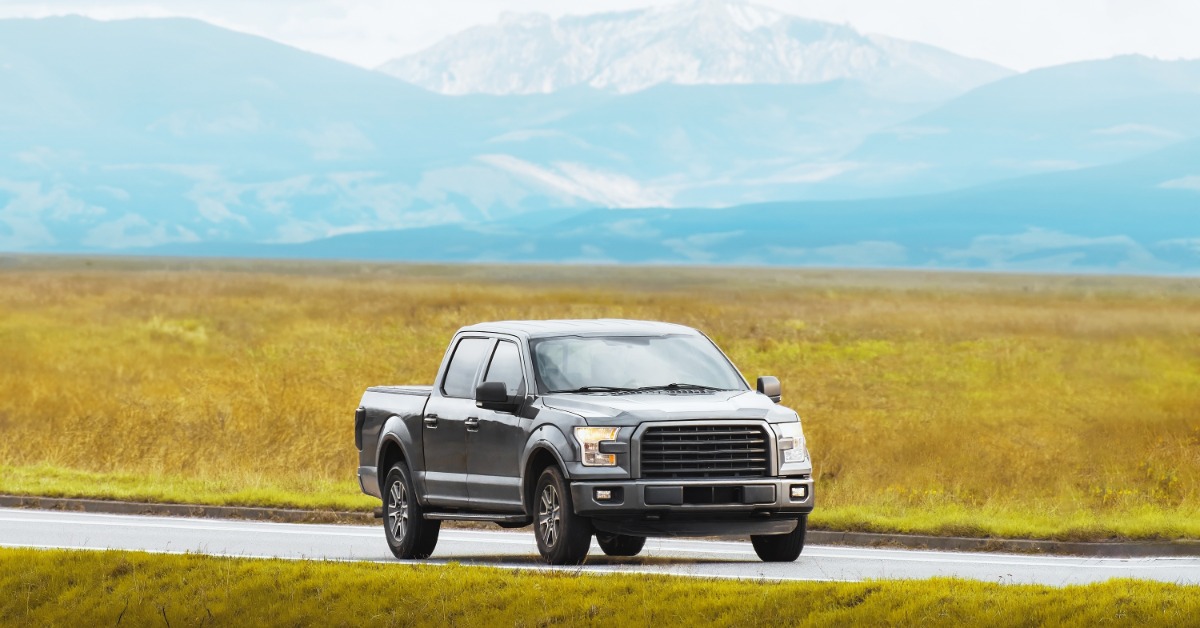 A black pickup truck traveling on a paved road beside yellow grass with blue mountains and cloudy sky beyond.