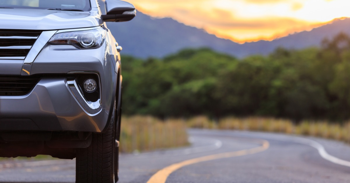 A silver SUV is driving on a lined asphalt road. Green trees, mountains, and a sunset are in the background.