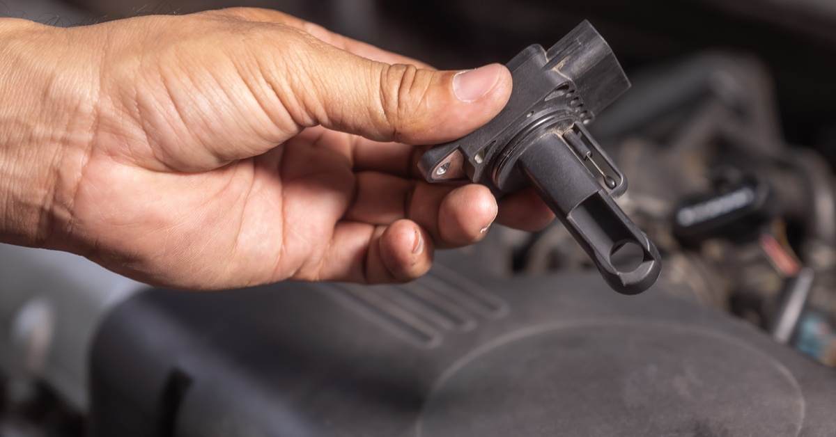A close-up of a hand holding a black mass air flow sensor for a car above an open engine compartment. A close-up of a hand holding a black mass air flow sensor for a car above an open engine compartment.