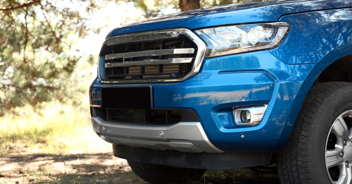 A blue pickup truck front end parked on dirt and grass, with a chrome grille, headlight, fog light, and trees.