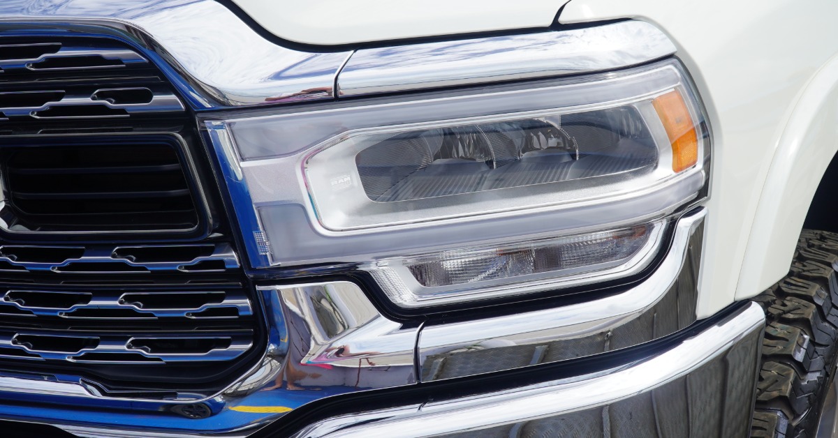 A close-up of a white pickup truck front end with a chrome grille, headlight unit, amber side marker, and tire.