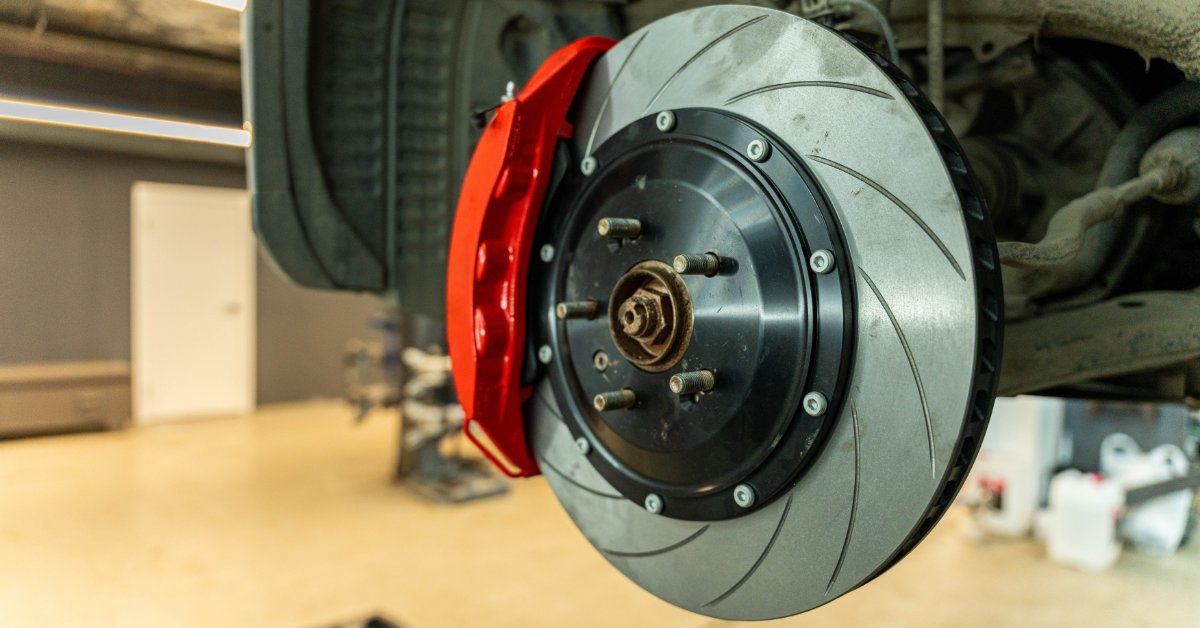 A red brake caliper beside a grooved ventilated silver rotor mounted on a raised vehicle in an auto repair bay.