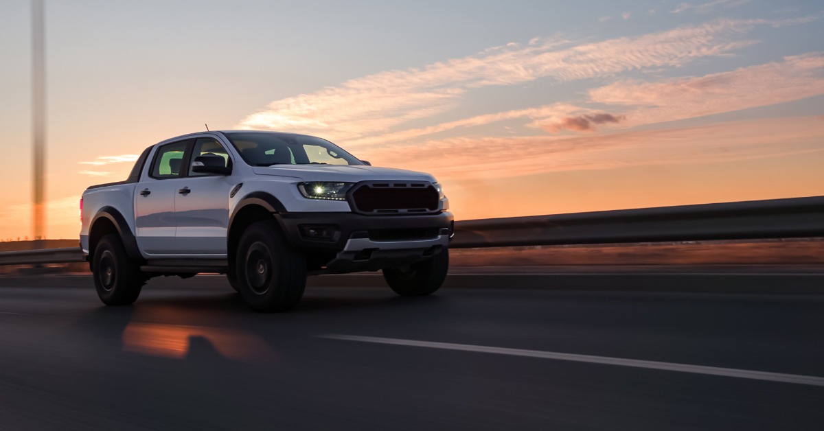 A white four-door pickup truck with black wheels and lit headlights drives along the highway beneath a pink sunset.