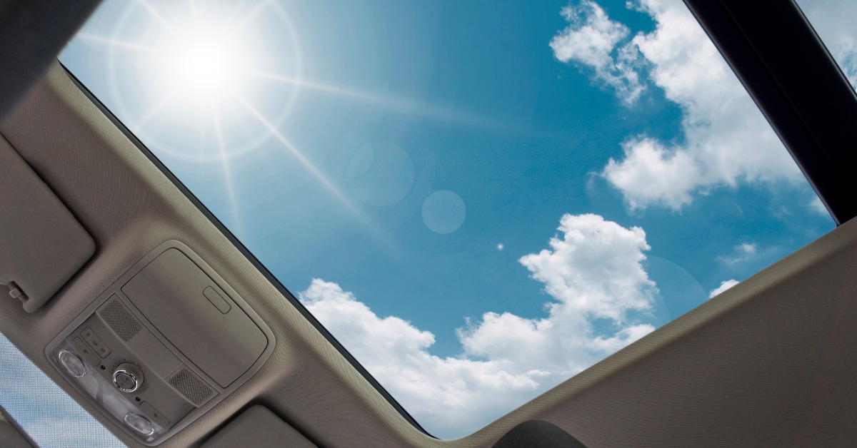 A large car sunroof surrounded by gray interior trim, showing bright sunlight and white clouds against a blue sky.