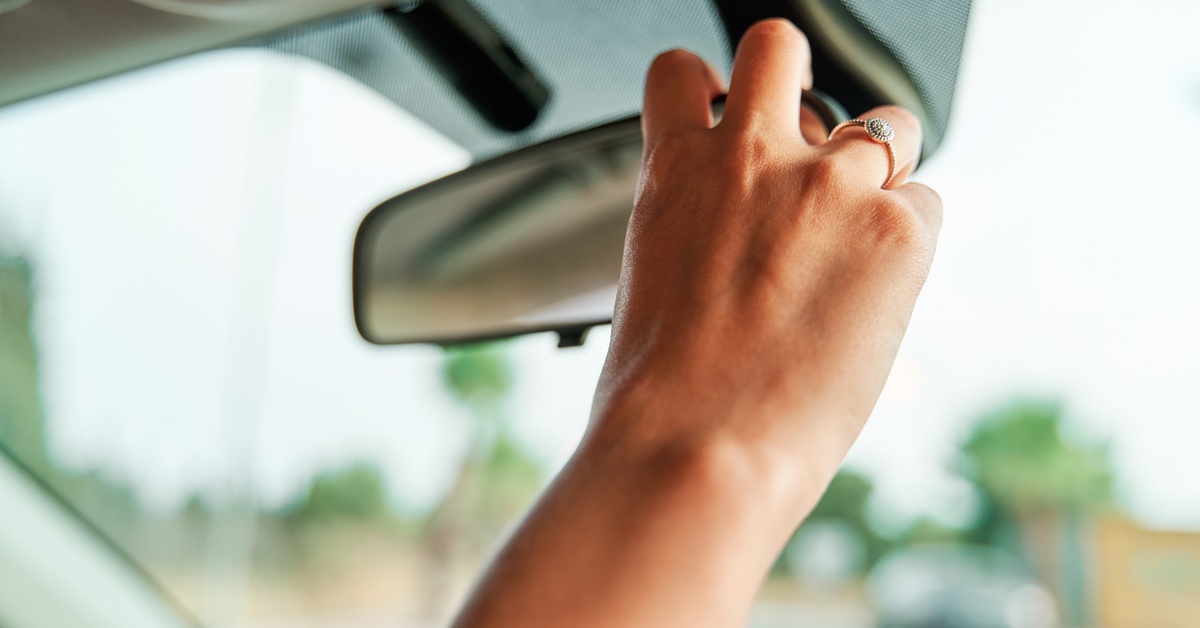 A person adjusting their rearview mirror with their right hand. The rearview mirror is mounted to the windshield.