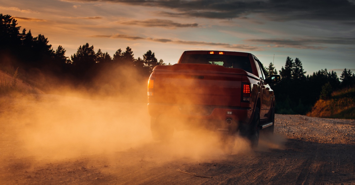 A red pickup truck driving through a desert terrain. There is a cloud of dust and dirt forming behind the vehicle.