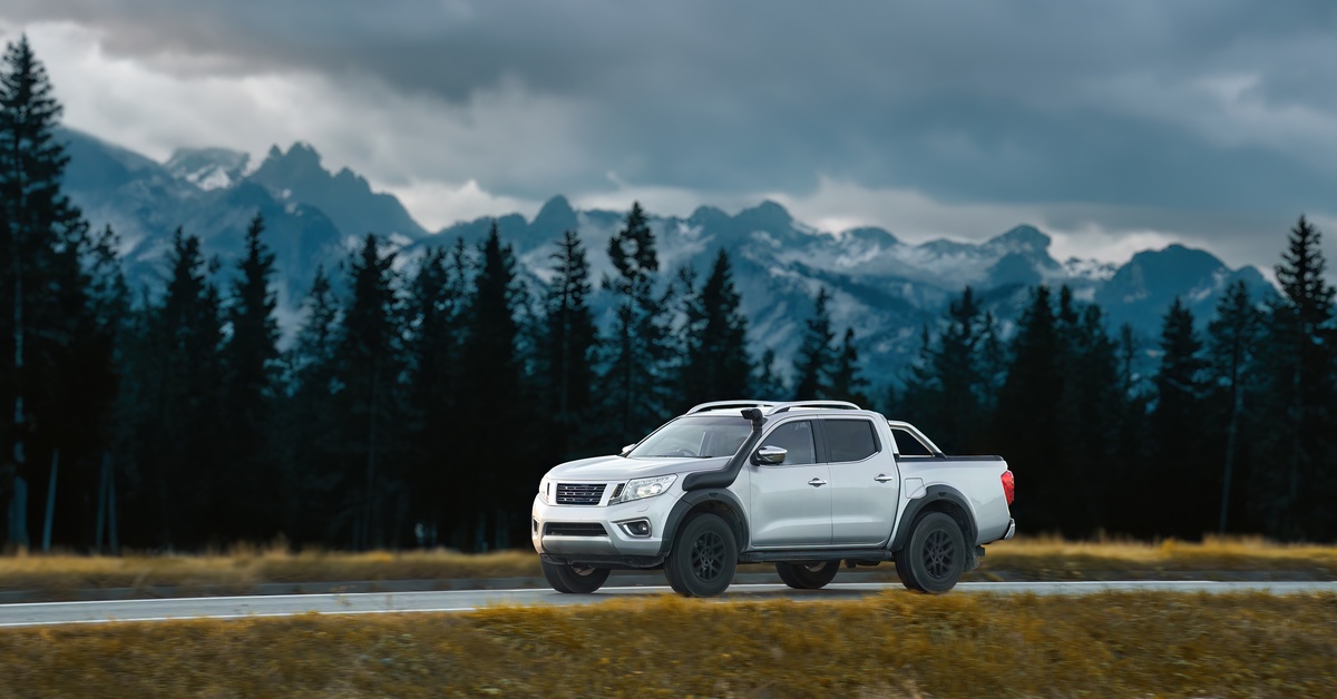 A white pickup truck driving down an empty highway. There are tall trees and snowy mountains surrounding the road.