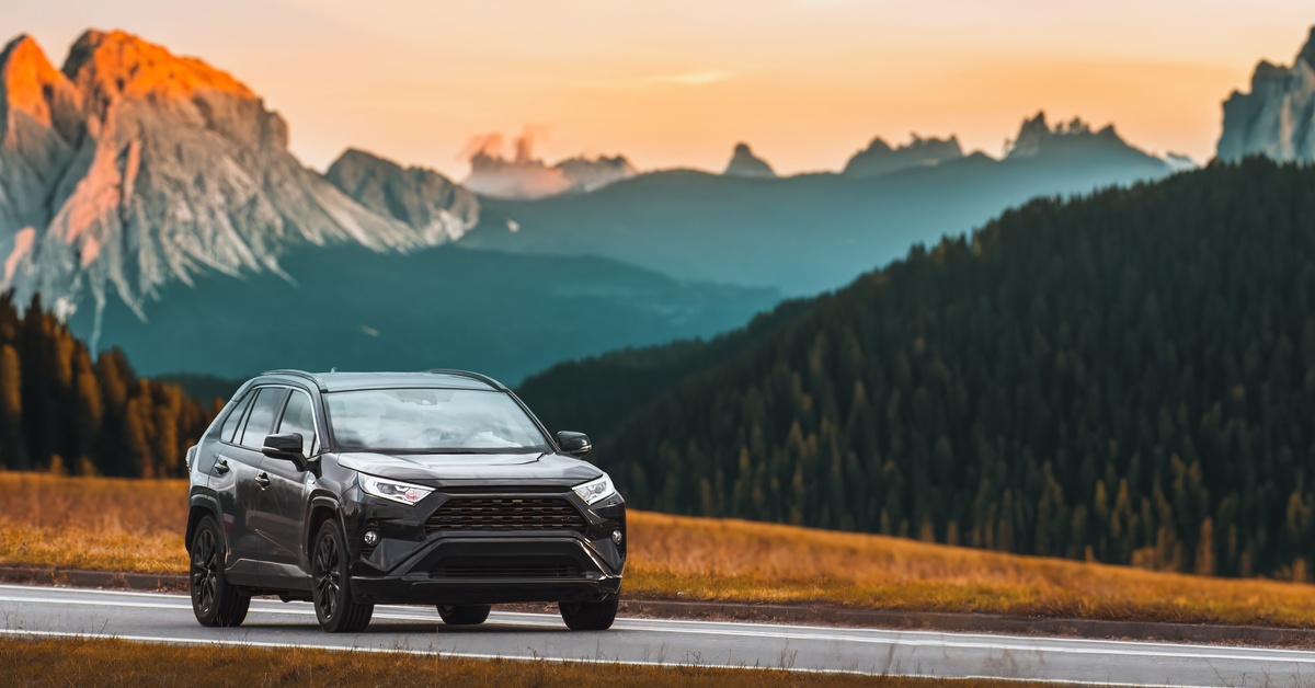 A black SUV driving down a one-lane highway. Tall mountain ranges and forests are behind the vehicle.