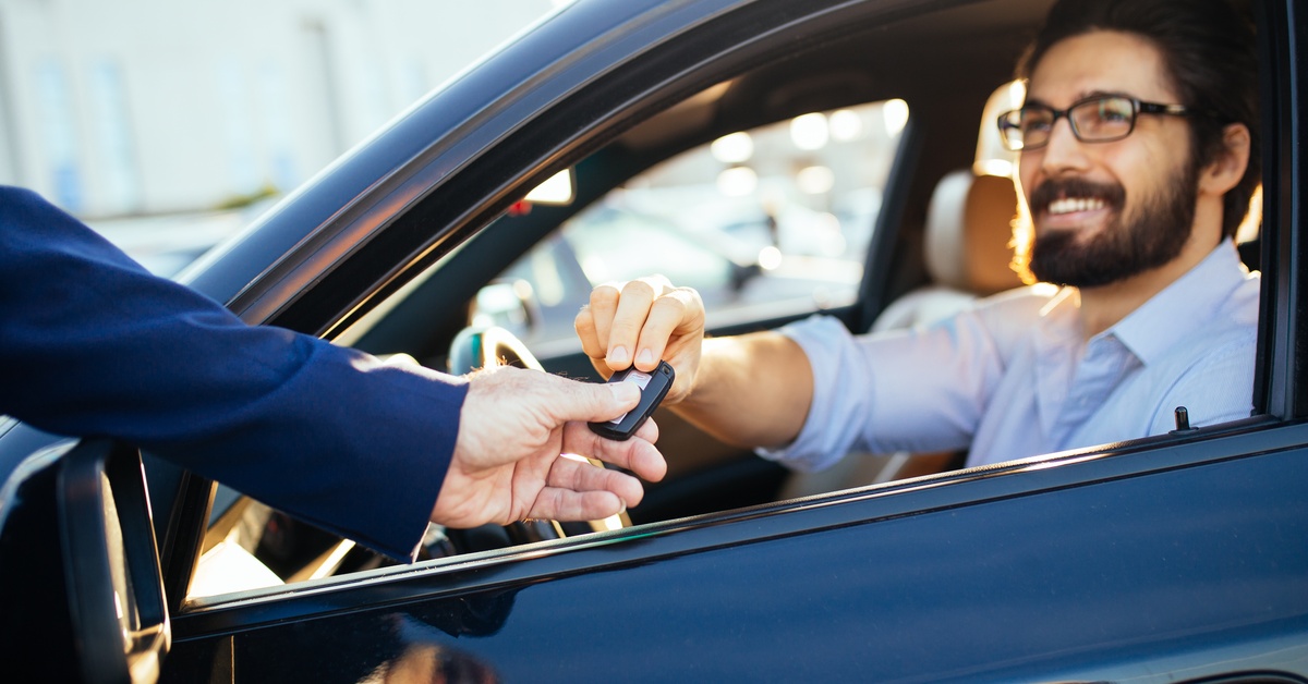 A man sitting in the driver’s seat of a car, receiving the electronic car keys from another person outside of the vehicle.
