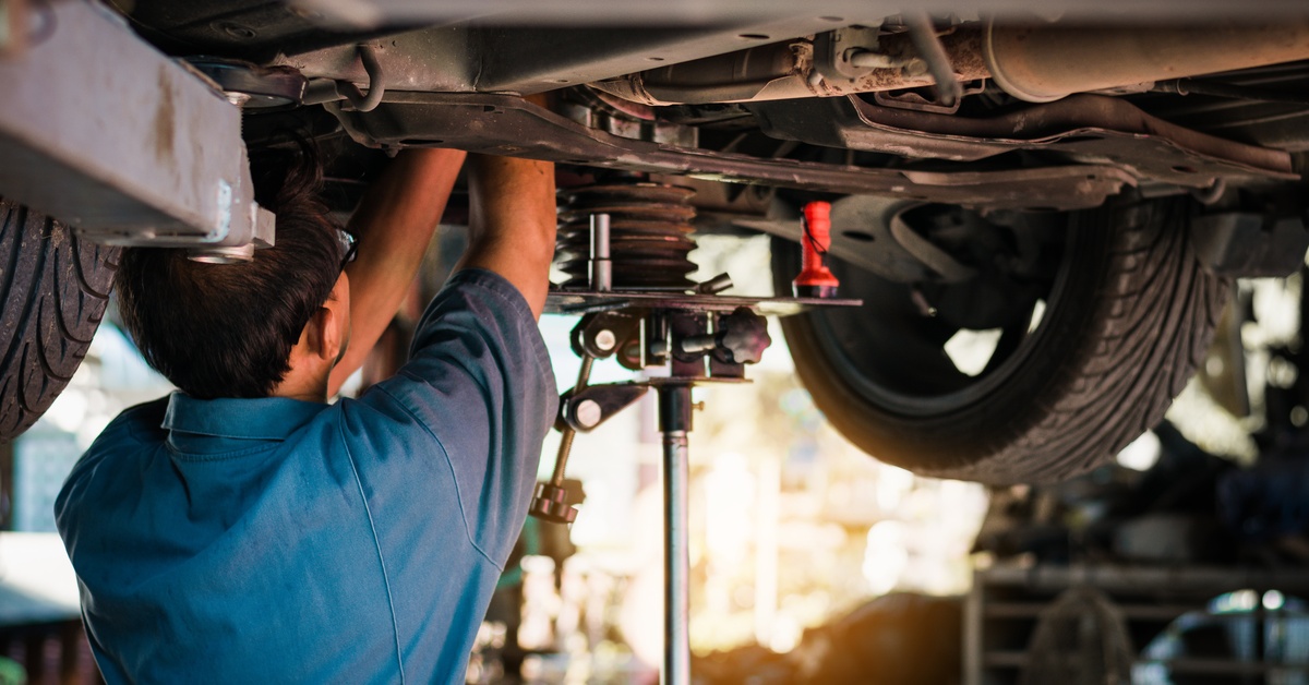 A mechanic conducting repairs to a vehicle in an auto shop from underneath the car. He is wearing a blue denim shirt.