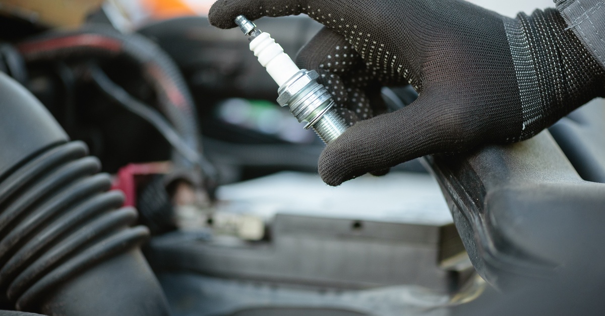 A close-up of a person wearing black gloves holds a spark plug. A vehicle under repair is in the background.