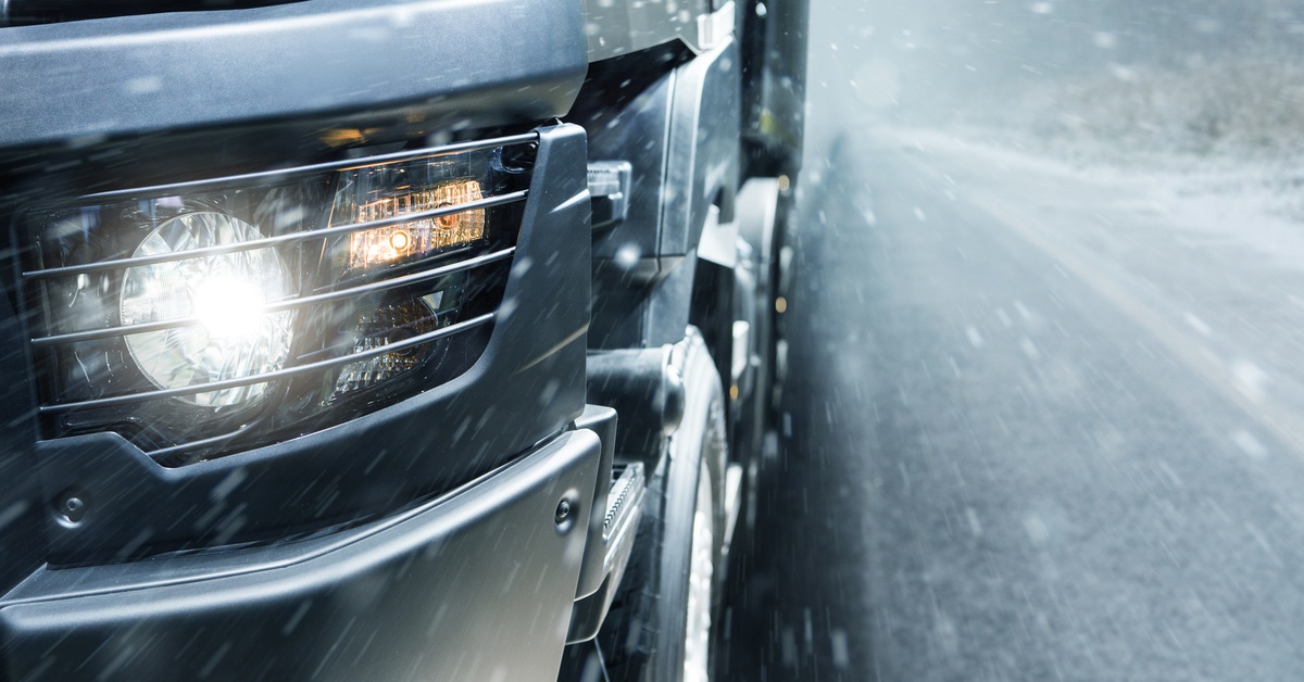An extreme close-up of the headlight of a silver truck. The vehicle is driving down a road as snow falls.