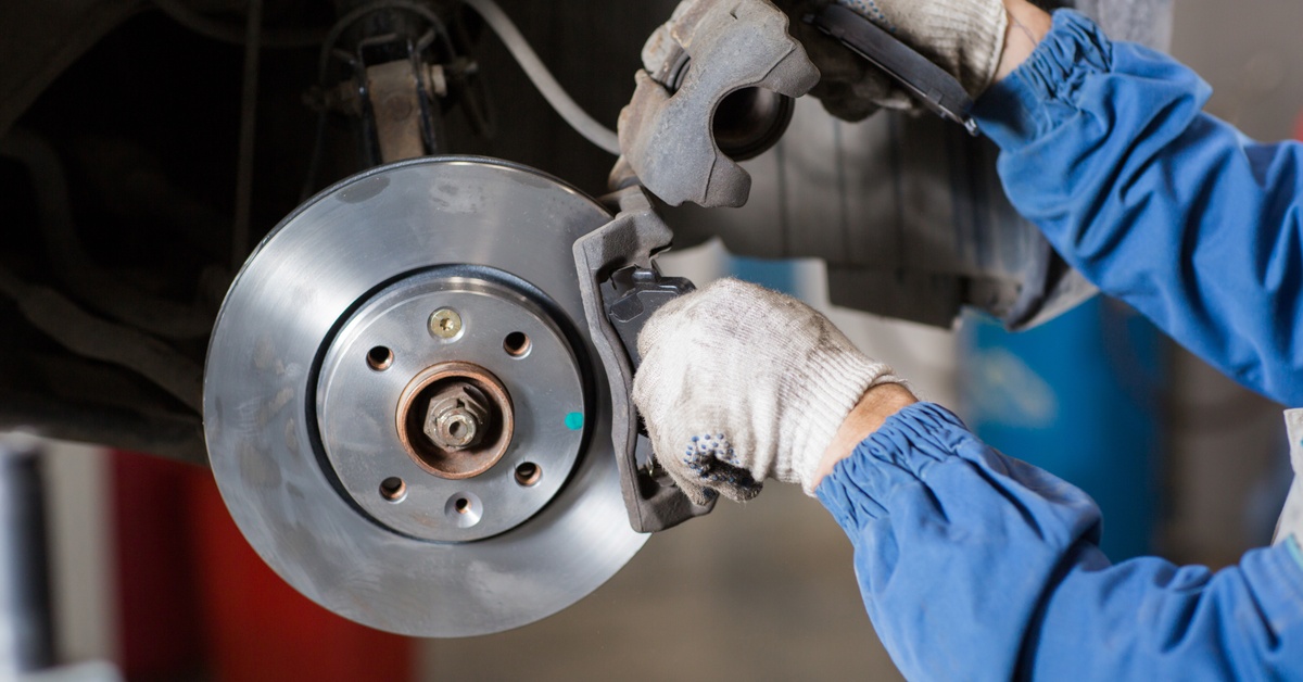 A person adjusts a brake caliper and pad near a shiny disc rotor. Tools and car parts are blurred in the background.