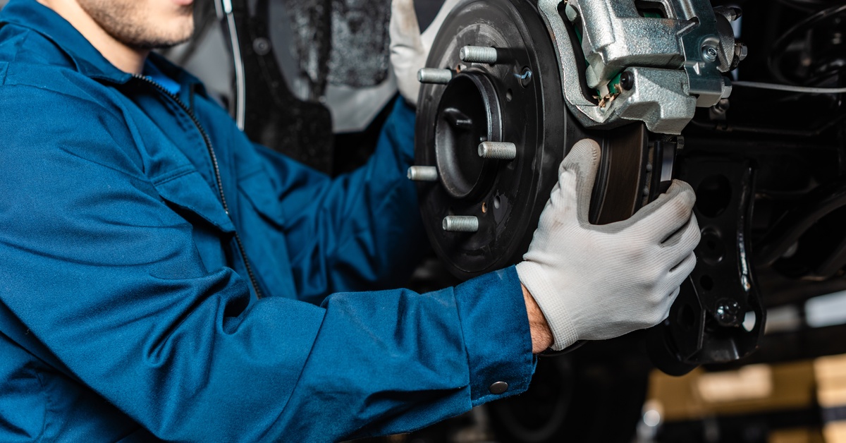 A mechanic in blue overalls works on a car's brake system, holding a brake pad near the rotor and caliper in a garage.