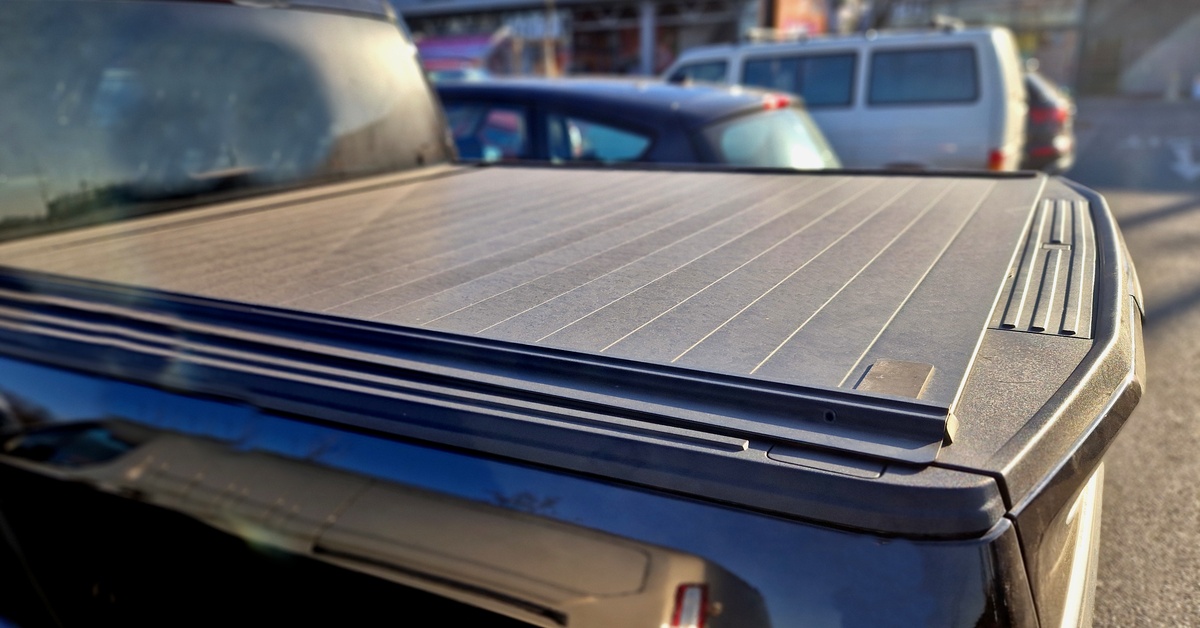 A truck is parked in a parking lot in front of shops. The truck has a black tonneau cover over the bed.