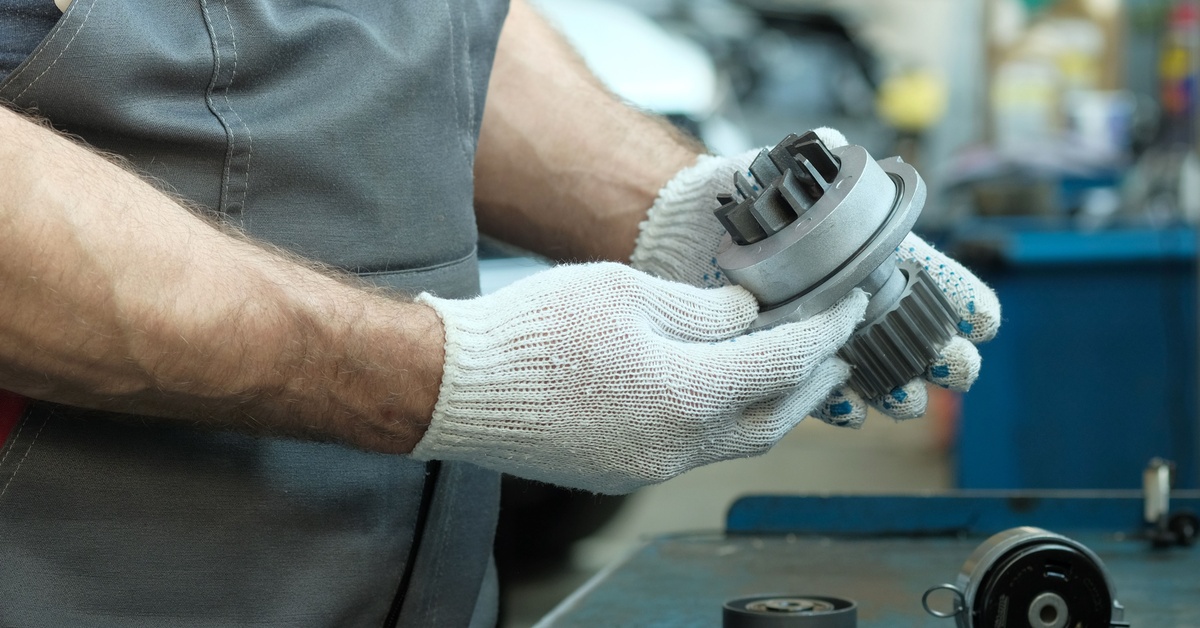 A mechanic wearing gray overalls and white gloves holds a metal, automotive water pump in a workshop.