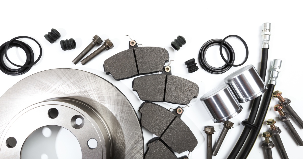 An assortment of brake parts are lying flat on a white surface. The parts include brake pads, cylinders, and brake hoses.
