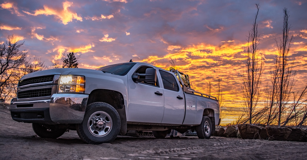 A white four-door truck is parked on a dirt road above a cliff as the sun sets. The sky is orange and cloudy. A white four-door truck is parked on a dirt road above a cliff as the sun sets. The sky is orange and cloudy.