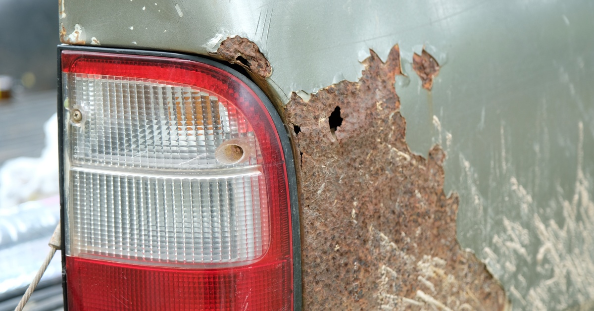 A red and clear tail light beside a large rusted hole and peeling green paint on the rear corner of a pickup truck. A red and clear tail light beside a large rusted hole and peeling green paint on the rear corner of a pickup truck.