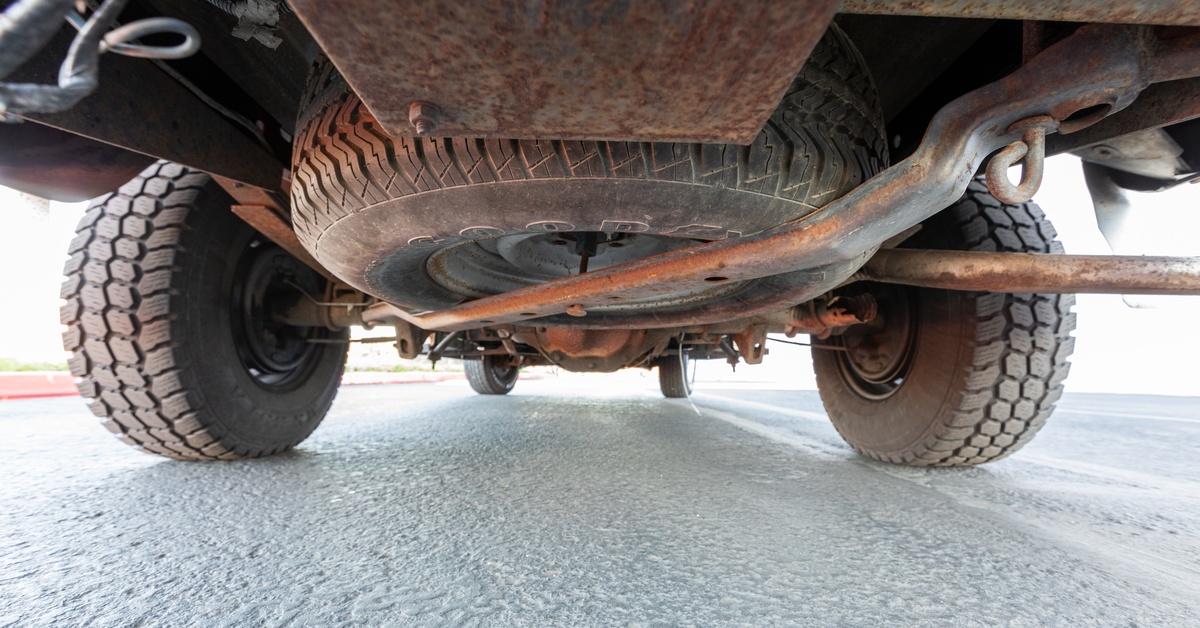 A rusty underside of a lifted pickup truck showing large tires, a spare tire, axle parts, and metal suspension components. A rusty underside of a lifted pickup truck showing large tires, a spare tire, axle parts, and metal suspension components.